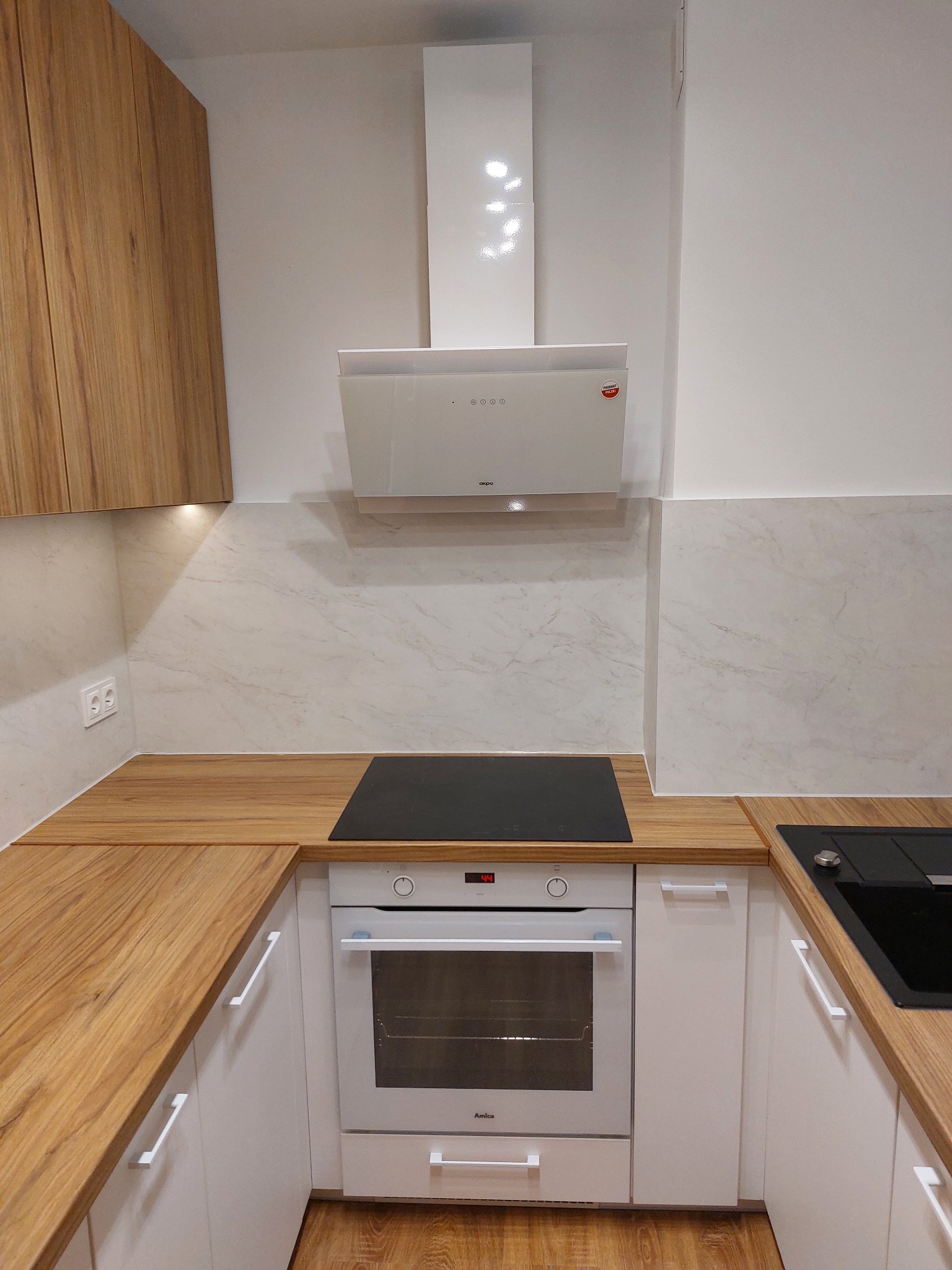 Nowoczesna zabudowa kuchenna z jasnymi frontami, drewnianym blatem i białym okapem, płyta indukcyjna wkomponowana w blat, piekarnik marki Amica.