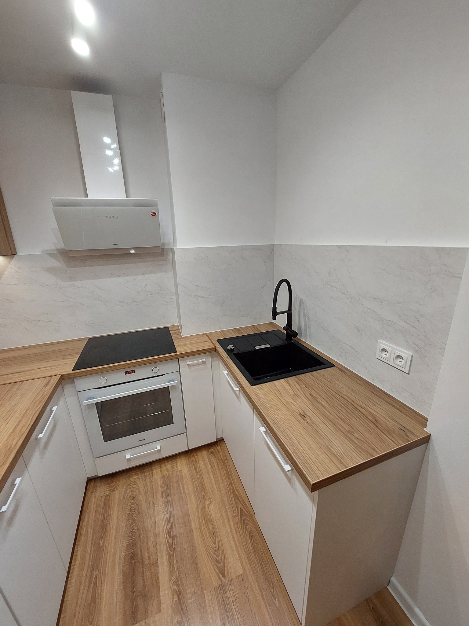 Nowoczesna kuchnia na wymiar w Kostrzynie: białe szafki z drewnianym blatem, czarny zlew z wysoką baterią i płyta indukcyjna pod białym okapem.