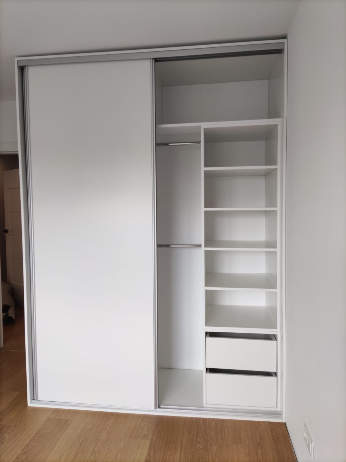 Biała szafa wnękowa z przesuwnymi drzwiami i otwartą częścią z półkami oraz dwoma szufladami, widoczna na tle jasnych ścian i drewnianej podłogi.