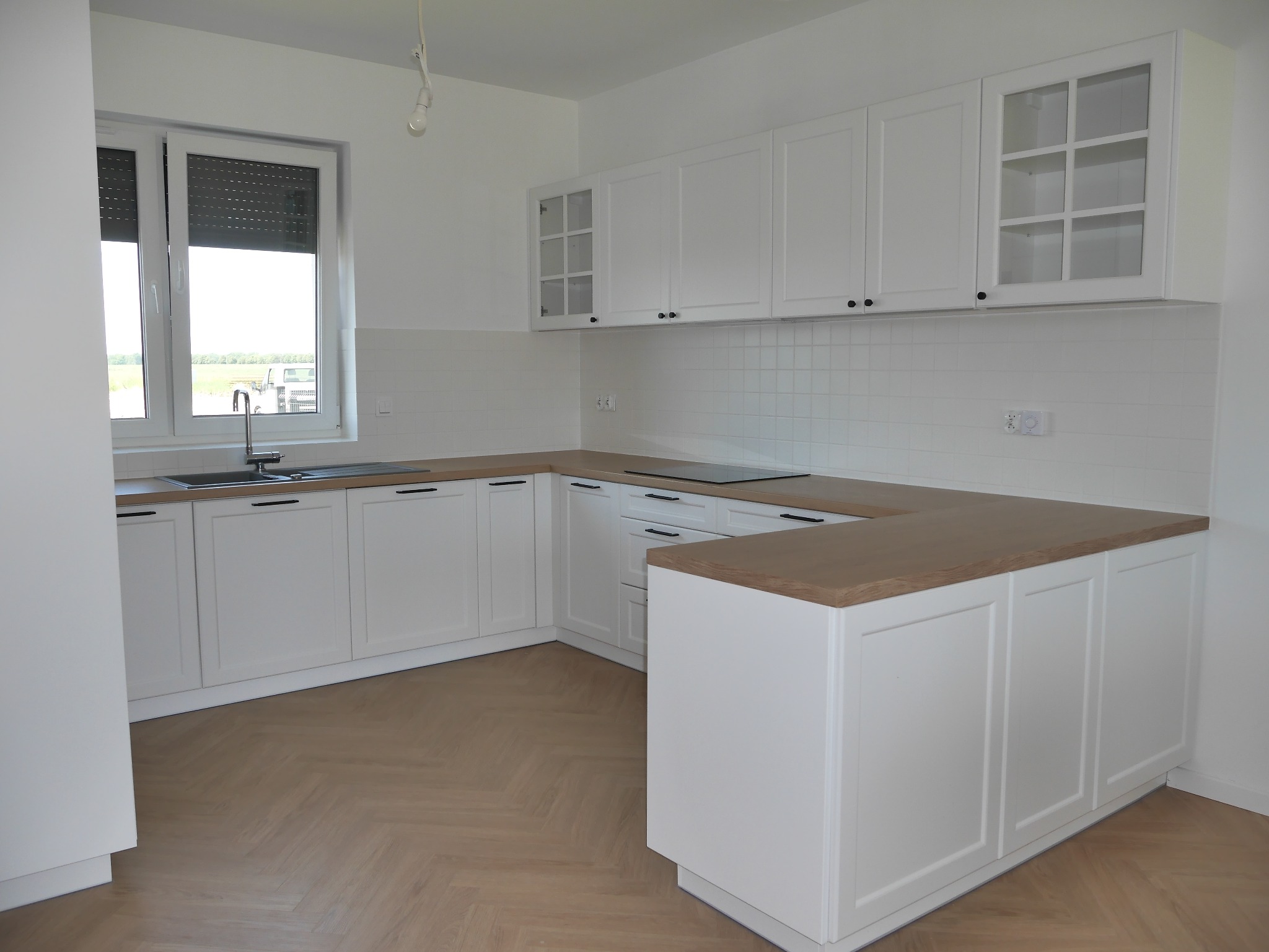 Jasna kuchnia z białymi szafkami i drewnianym blatem, widok na okno z żaluzjami. Podłoga wyłożona parkietem w jodełkę.