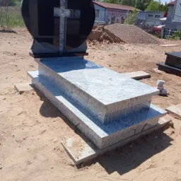 Granitowy pomnik z czarnym nagłówkiem w kształcie serca z jasnym krzyżem, wykonany w Ostródzie, ustawiony na piaszczystym podłożu.