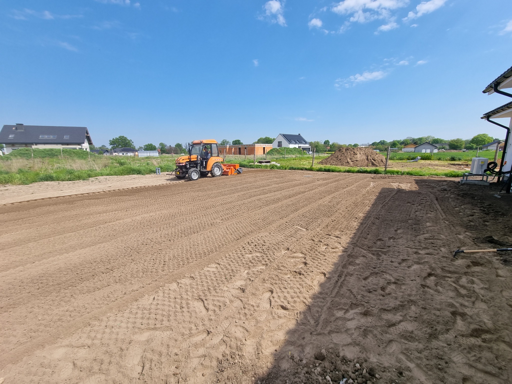 Pomarańczowy ciągnik z glebogryzarką przygotowuje pole pod zasiew, widoczne świeżo spulchnione bruzdy w ziemi, w tle zabudowa jednorodzinna i błękitne niebo.