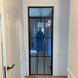 BART MEBLE - Wąski korytarz z białą boazerią i czarną, przeszkloną ścianką działową z metalowymi szprosami, odbicie osoby robiącej zdjęcie.