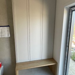 BART MEBLE - Wysoka, zabudowana szafa z trzema frontami w jasnym odcieniu szarości, z drewnianym siedziskiem u dołu, umieszczona we wnęce obok okna. Na ścianie widoczne plany techniczne.