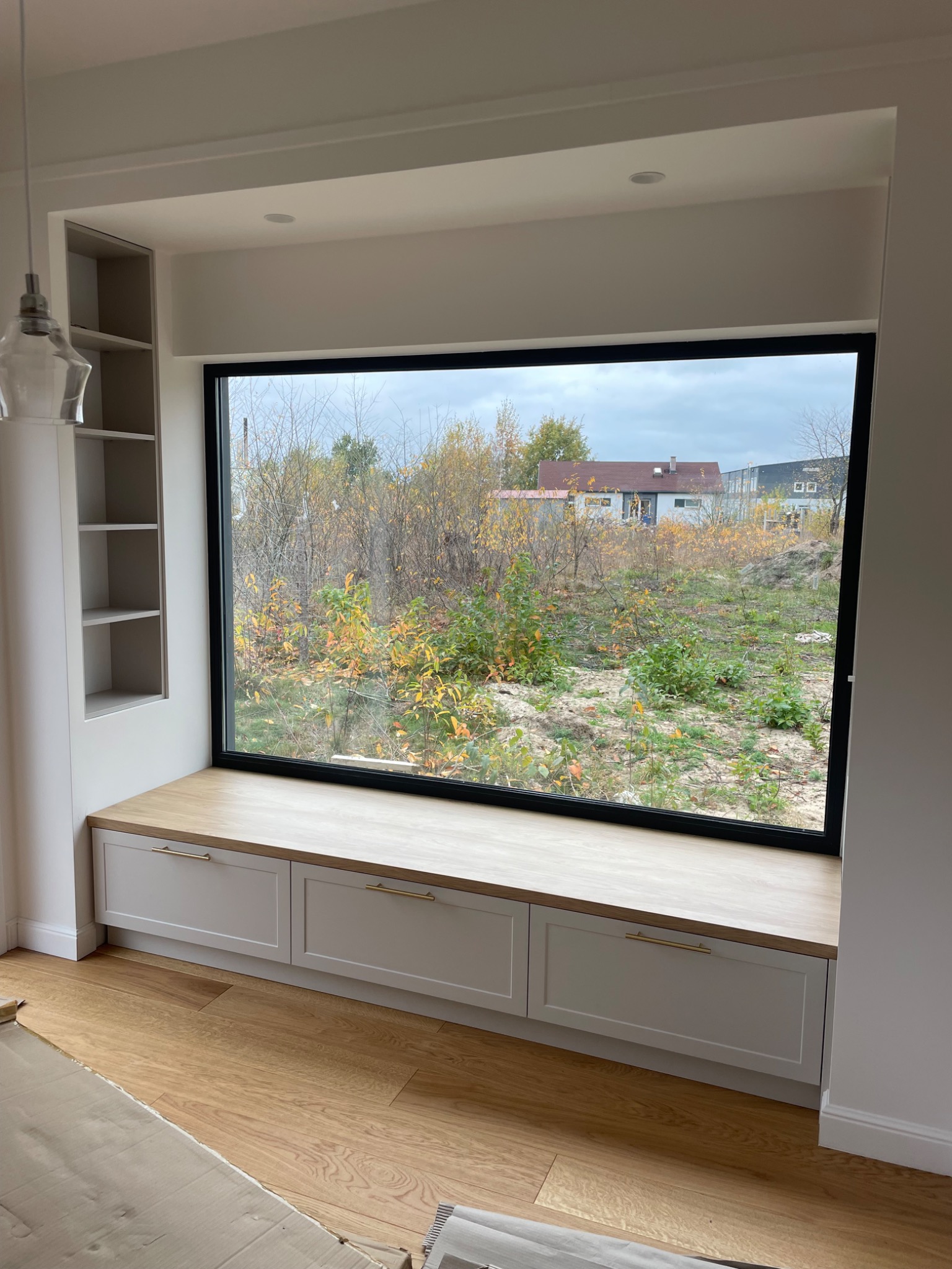 Wnęka okienna z siedziskiem i szafkami w kolorze złamanej bieli, drewniany blat i widok na ogród zza okna.