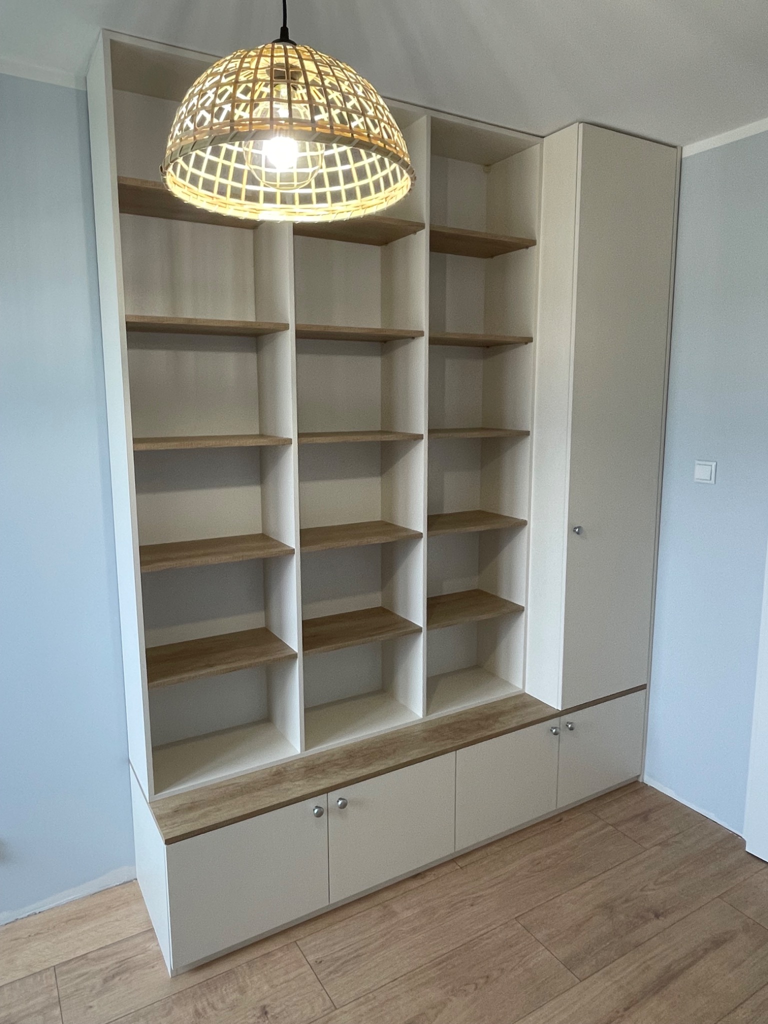 Biała szafa z otwartymi półkami z drewnianymi akcentami, wbudowana w ścianę, oświetlona dekoracyjną lampą z plecionki.