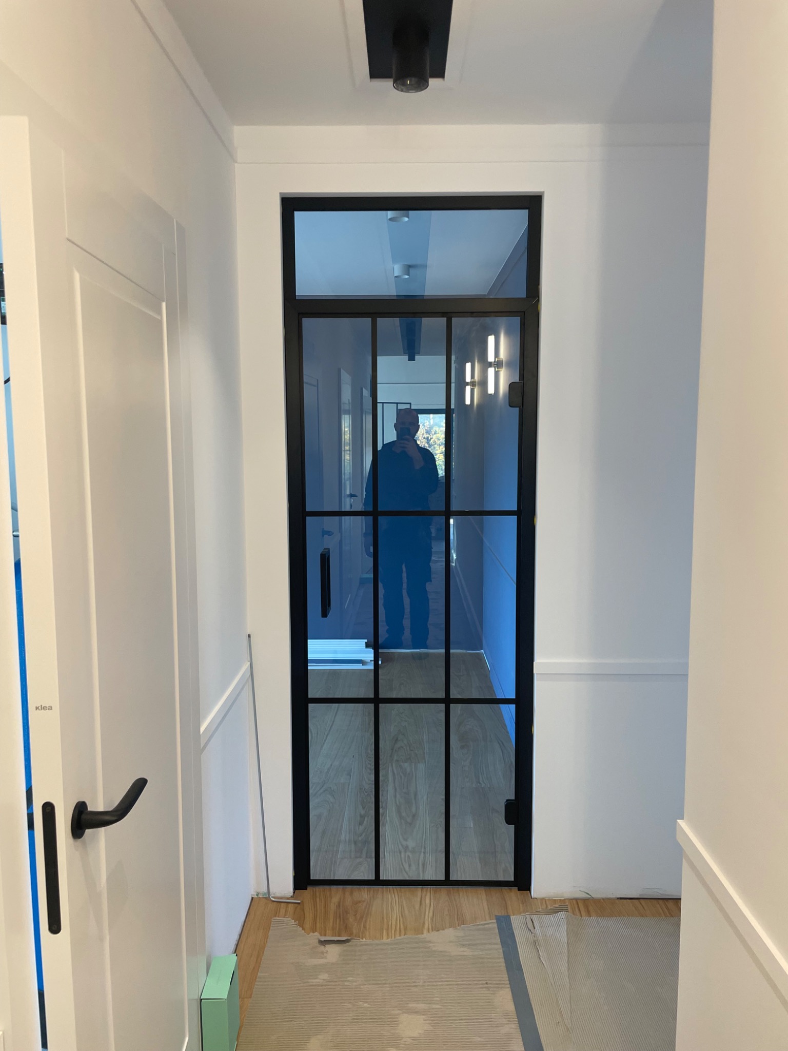 Wąski korytarz z białą boazerią i czarną, przeszkloną ścianką działową z metalowymi szprosami, odbicie osoby robiącej zdjęcie.