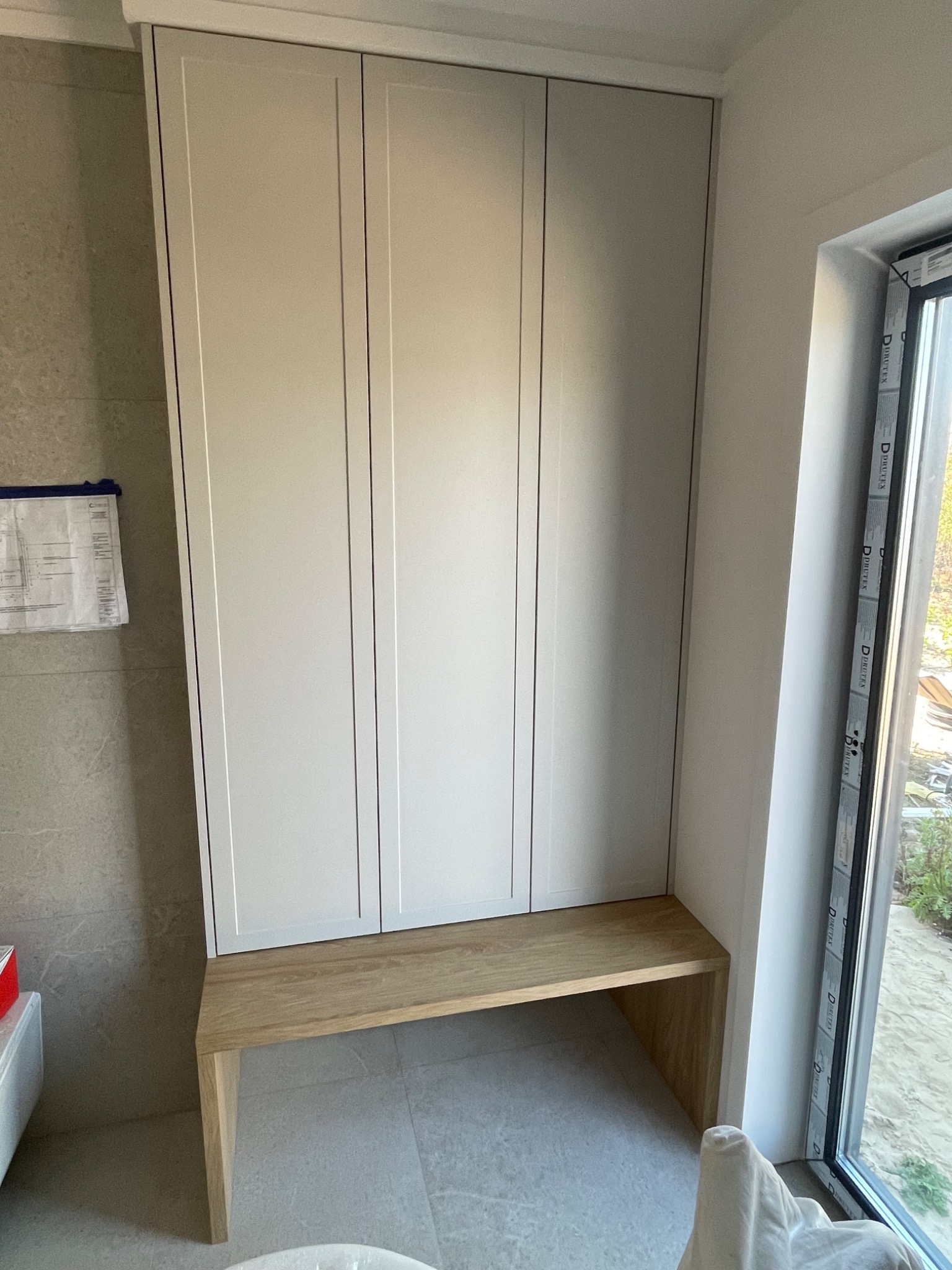 Wysoka, zabudowana szafa z trzema frontami w jasnym odcieniu szarości, z drewnianym siedziskiem u dołu, umieszczona we wnęce obok okna. Na ścianie widoczne plany techniczne.