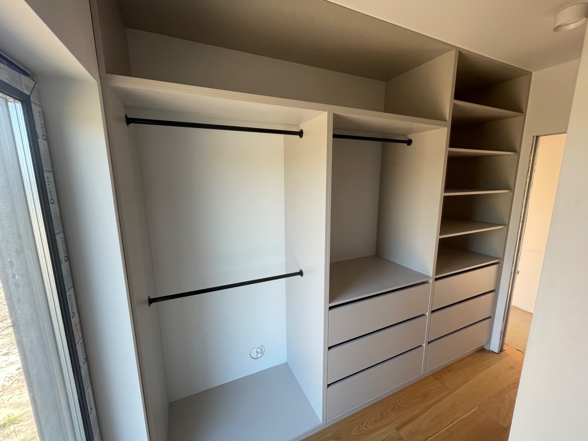 Wnętrze szafy wnękowej z szufladami i drążkami na ubrania, widok z perspektywy wnętrza pomieszczenia, obok okno z widoczną ramą.