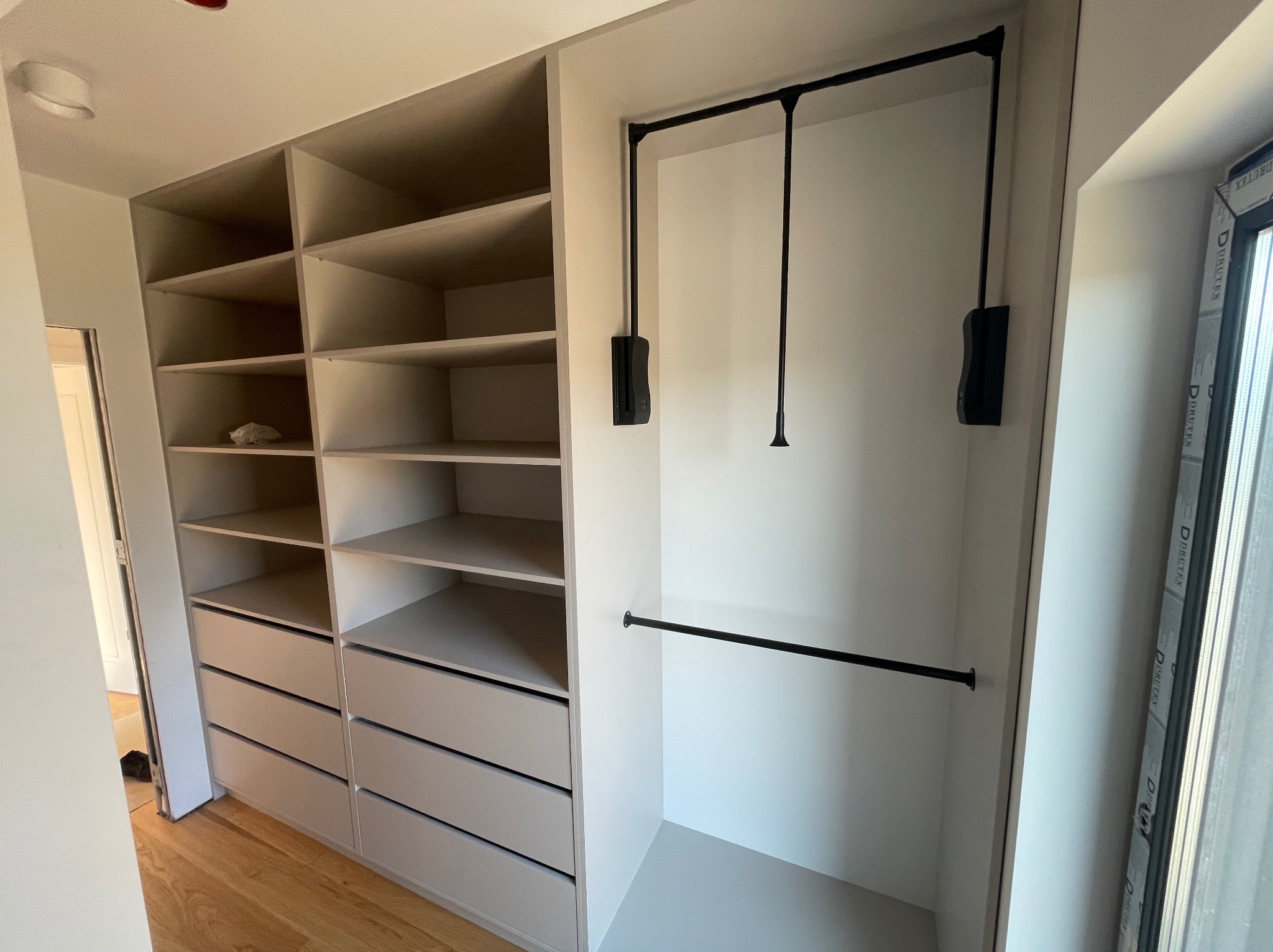 Wnętrze szafy wnękowej z półkami, szufladami i drążkiem na ubrania, widoczny mechanizm opuszczania drążka, jasna kolorystyka, drewniana podłoga.