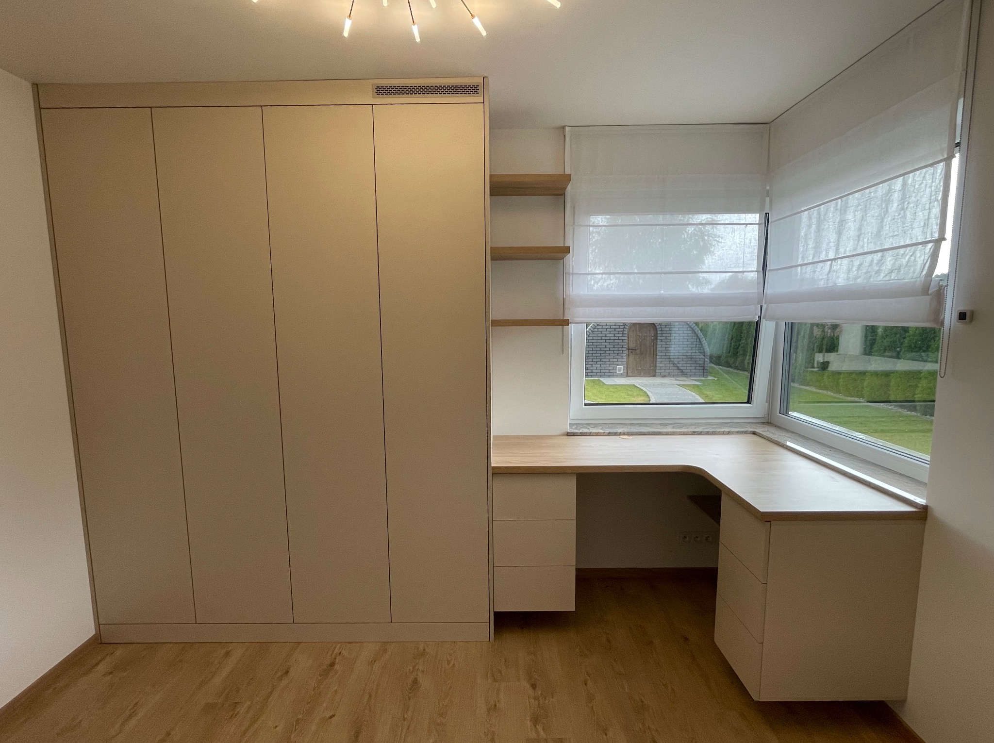 Beżowa szafa wnękowa z biurkiem pod oknem, drewniane półki i rolety rzymskie. Jasne wnętrze z parkietem. Minimalistyczny design, zabudowa na wymiar.