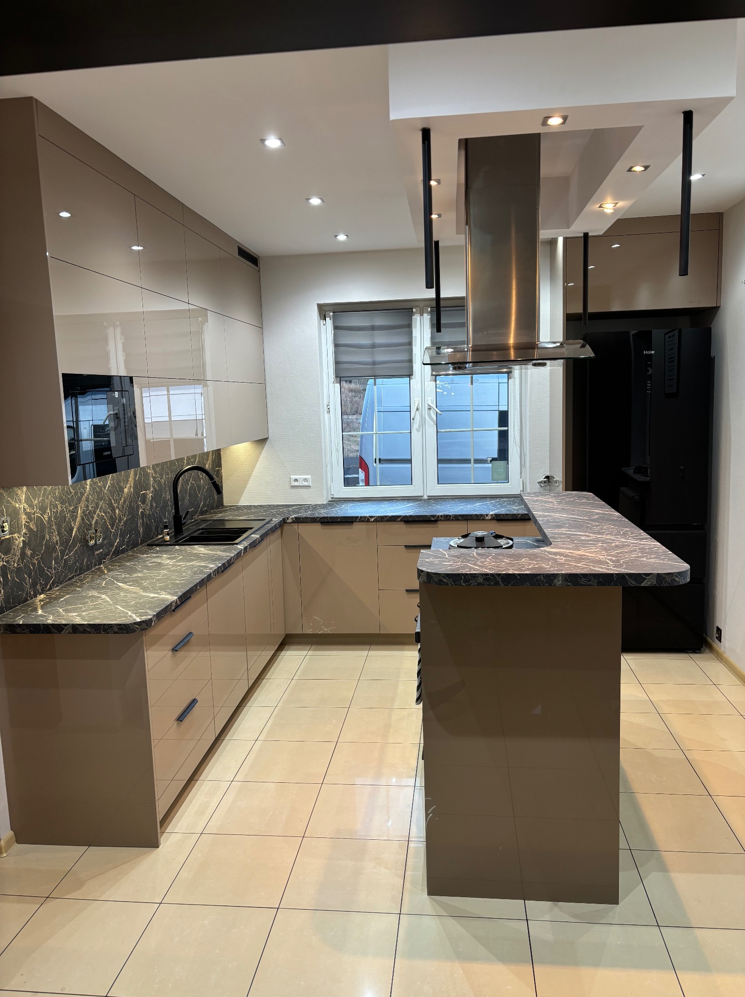 Beżowa kuchnia z wyspą i blatem imitującym marmur. Nowoczesny design z błyszczącymi frontami i czarnymi akcentami. Widoczny okap i czarna lodówka.