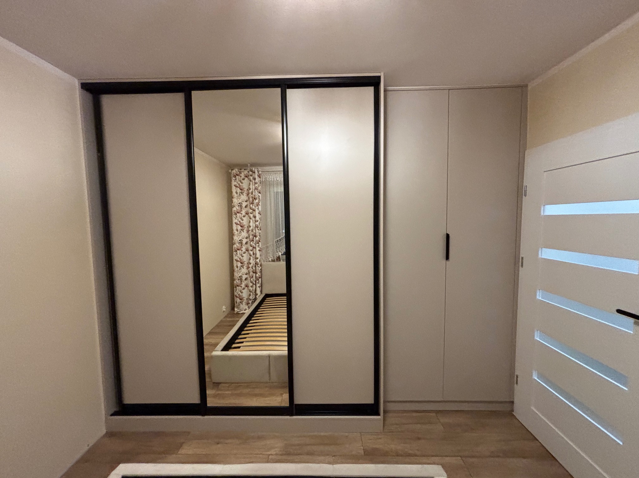 Jasna szafa wnękowa z przesuwnymi drzwiami, z lustrem odbijającym wnętrze sypialni. Obok szafa z długimi uchwytami i białe drzwi z poziomymi przeszkleniami.