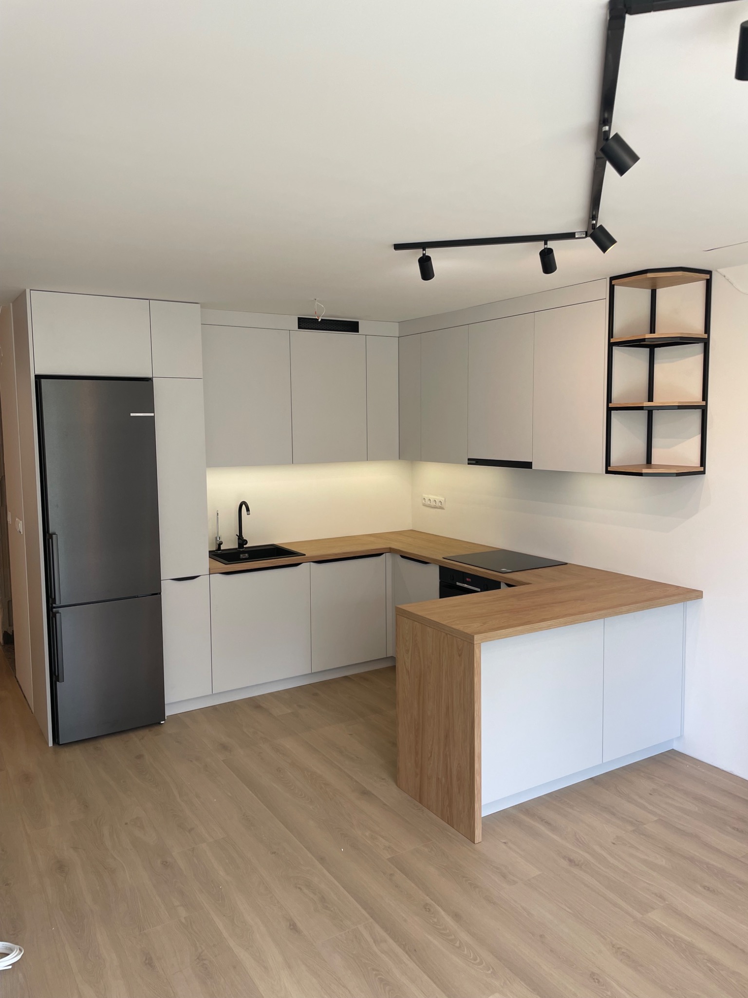 Nowoczesna kuchnia w kształcie litery U z drewnianym blatem i białymi szafkami. Czarny zlew i bateria, oświetlenie podszafkowe LED, minimalistyczny design.