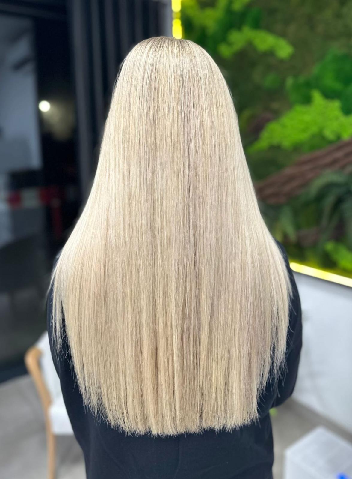 Długie, proste, platynowe włosy z delikatnym odrostem, widok z tyłu na tle dekoracji z roślin.
