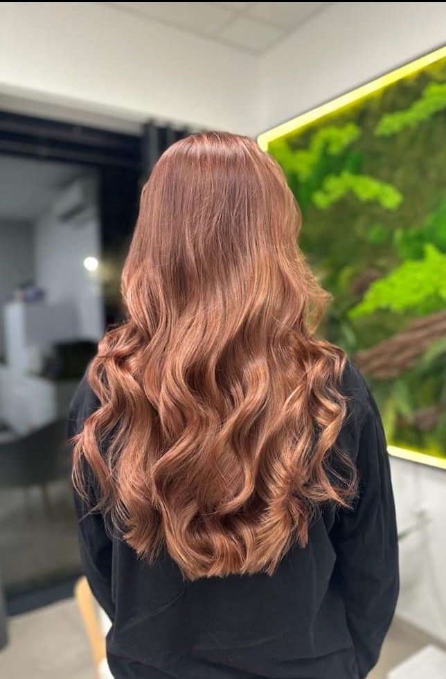 Długie, falowane włosy w odcieniach miedzianego blondu, z widocznymi refleksami, prezentowane na tle ściany z zielonym panelem dekoracyjnym w salonie fryzjerskim.