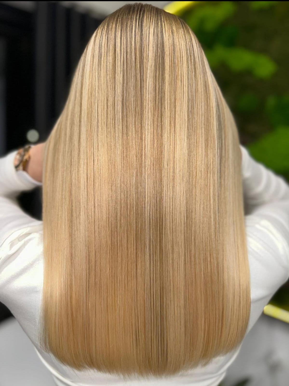 Proste, długie włosy blond z subtelnymi, jaśniejszymi pasemkami, widoczne od tyłu, z delikatnym połyskiem i idealnym wygładzeniem, na rozmytym tle z zielenią i czernią.