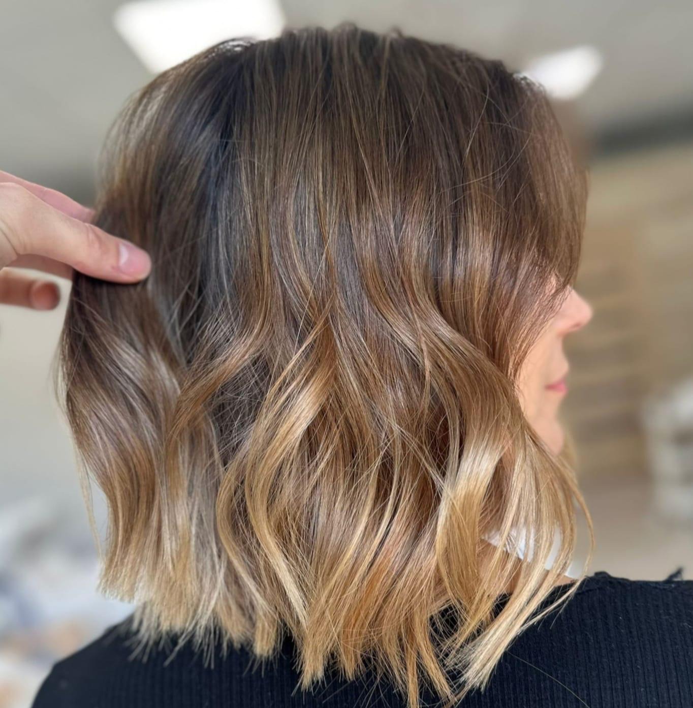 Włosy po koloryzacji sombre z delikatnymi falami, dłoń stylisty unosi pasmo włosów, prezentując przejścia kolorystyczne.