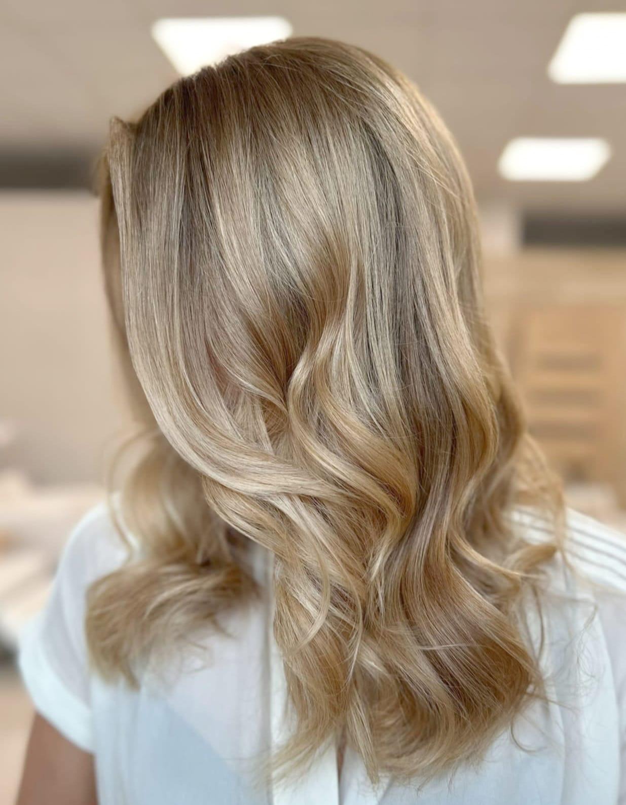 Blond włosy z delikatnymi falami, świeżo po koloryzacji i stylizacji, widoczne refleksy i przejścia tonalne, biała bluzka.