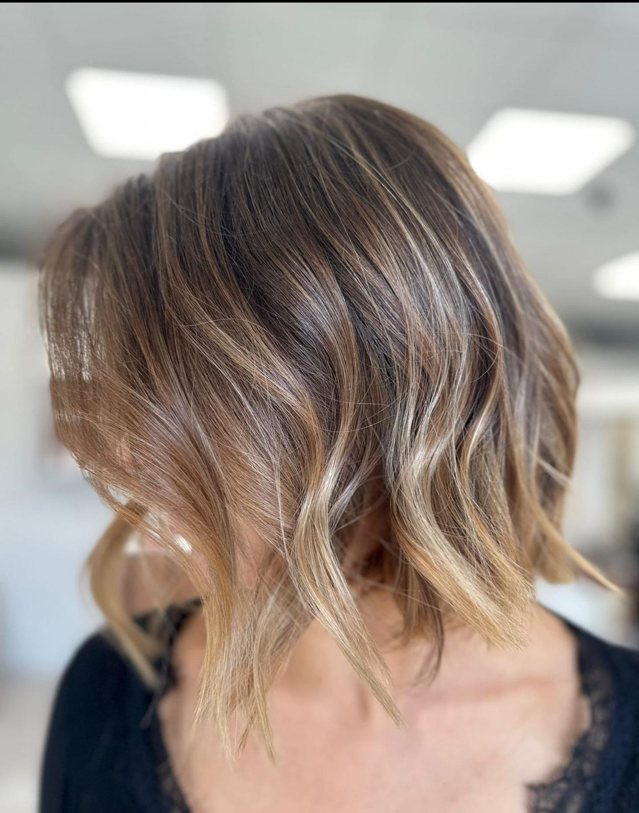 Krótkie, falowane włosy w odcieniach brązu i blondu, z subtelnymi refleksami, widoczne od tyłu, z delikatnym koronkowym wykończeniem bluzki.