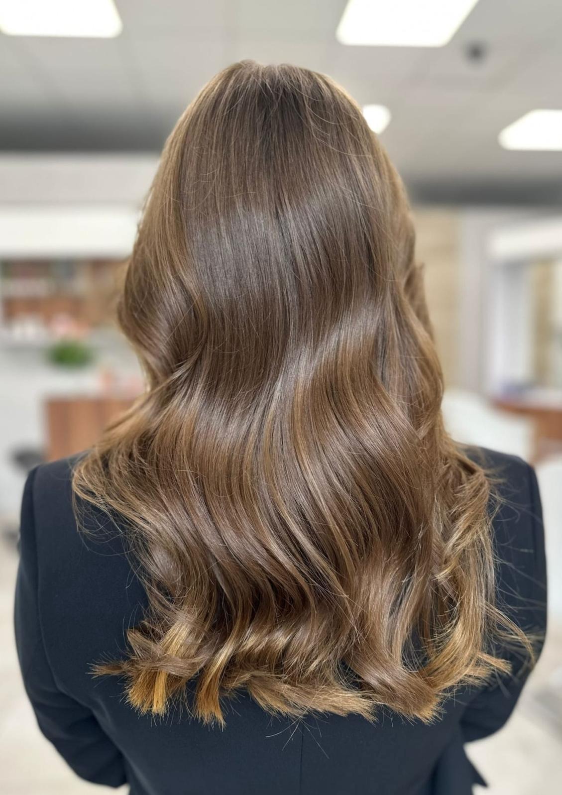 Średniej długości, brązowe włosy z delikatnymi falami i jaśniejszymi refleksami u dołu, widok z tyłu na tle jasnego wnętrza salonu.