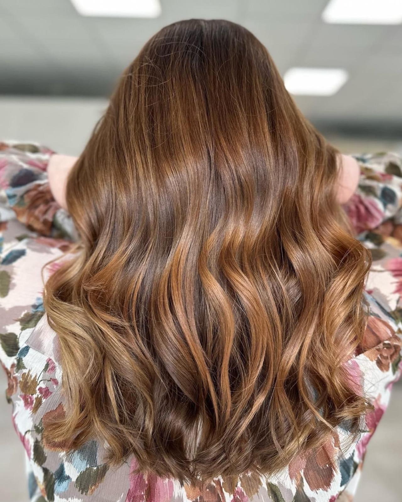 Falowane włosy w odcieniach brązu i blondu, widok z tyłu, delikatne refleksy, bluzka w kwiatowy wzór.