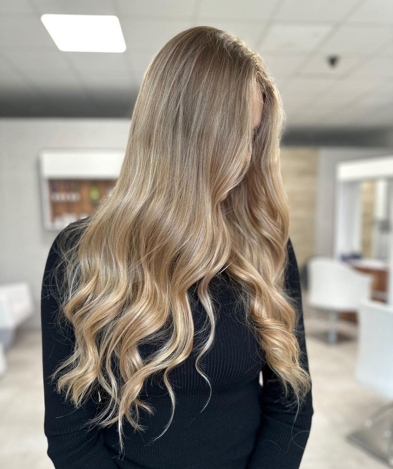 Blond włosy z delikatnymi falami, stylizacja wykonana w salonie fryzjerskim, widok z tyłu, lekko rozmyte tło.