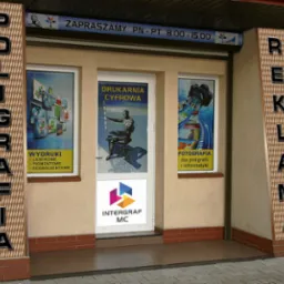 Wejście do drukarni cyfrowej Intergraf MC z reklamami wydruków laserowych, pigmentowych i ecosolventowych oraz fotografii dla poligrafii i informatyki. Na fasadzie pionowe szyldy z napisami...