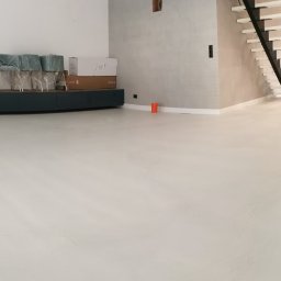 Żywica i kamień - Jasna, jednolita podłoga w przestronnym wnętrzu z nowoczesnymi schodami i wyspą kuchenną. Krzesła w folii ochronnej i kartony stoją pod ścianą.