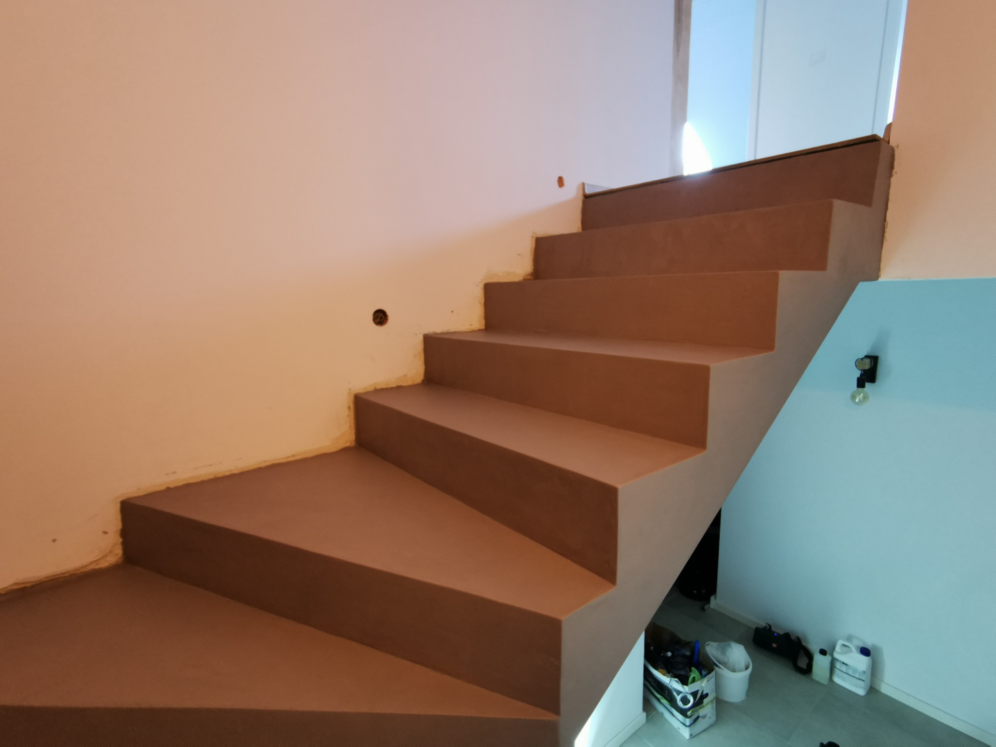 Schody wewnętrzne wykończone brązową masą, widoczne niedoskonałości na ścianie i materiały budowlane na podłodze wskazują na etap wykończeniowy.