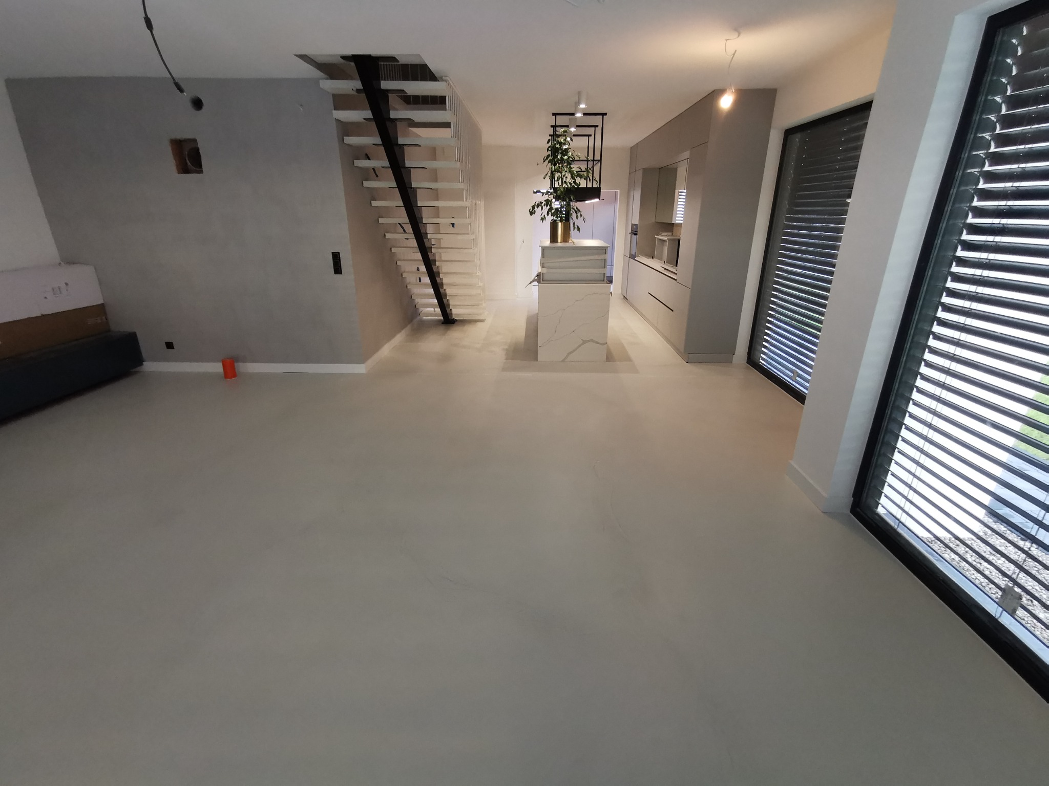 Jasna posadzka żywiczna w minimalistycznym wnętrzu z otwartą przestrzenią, schodami z czarnymi elementami i dużą wyspą kuchenną z blatem imitującym marmur.