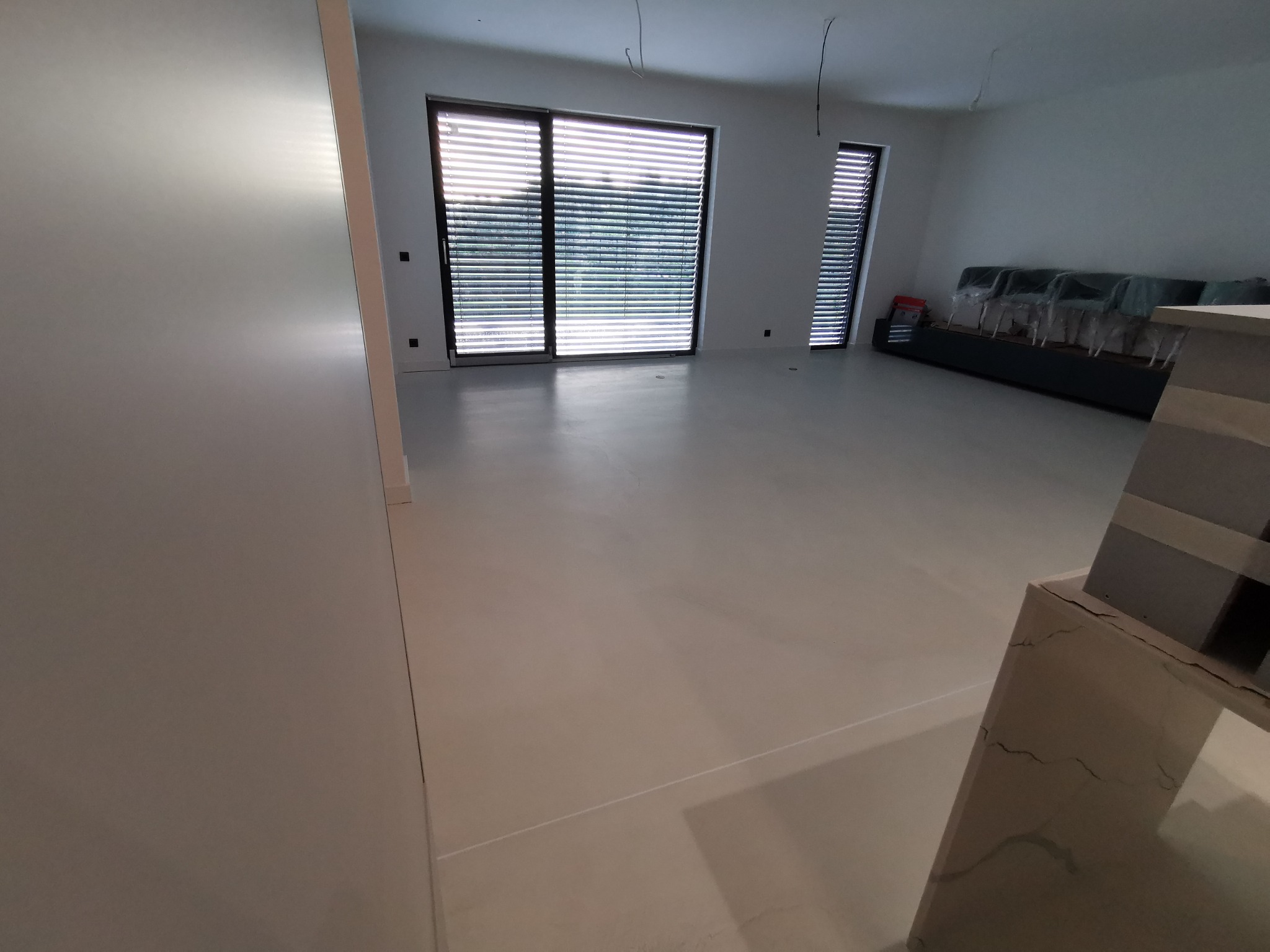 Jasnoszara, gładka podłoga w nowym, minimalistycznym wnętrzu z dużymi oknami z żaluzjami. W tle meble zabezpieczone folią.
