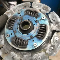 Zużyte sprzęgło samochodowe z widocznymi śladami przegrzania i zużycia, z niebieskawymi przebarwieniami termicznymi na tarczy, trzymane w rękawicy roboczej na tle warsztatu.