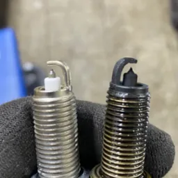 Porównanie nowej, srebrnej świecy zapłonowej z zużytą, czarną i zaolejoną świecą, trzymanych w dłoni w czarnej rękawicy roboczej.