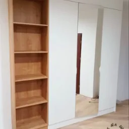 Biała szafa z lustrzanymi drzwiami i przylegającym drewnianym regałem z jasnego drewna, widoczny fragment podłogi i logo firmy w rogu.