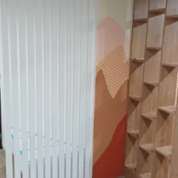 Biała ażurowa ścianka działowa z pionowych listew, przysłaniająca częściowo białe łóżko, obok drewniany regał z półkami w kształcie trójkątów i malowidło ścienne w geometryczne góry.