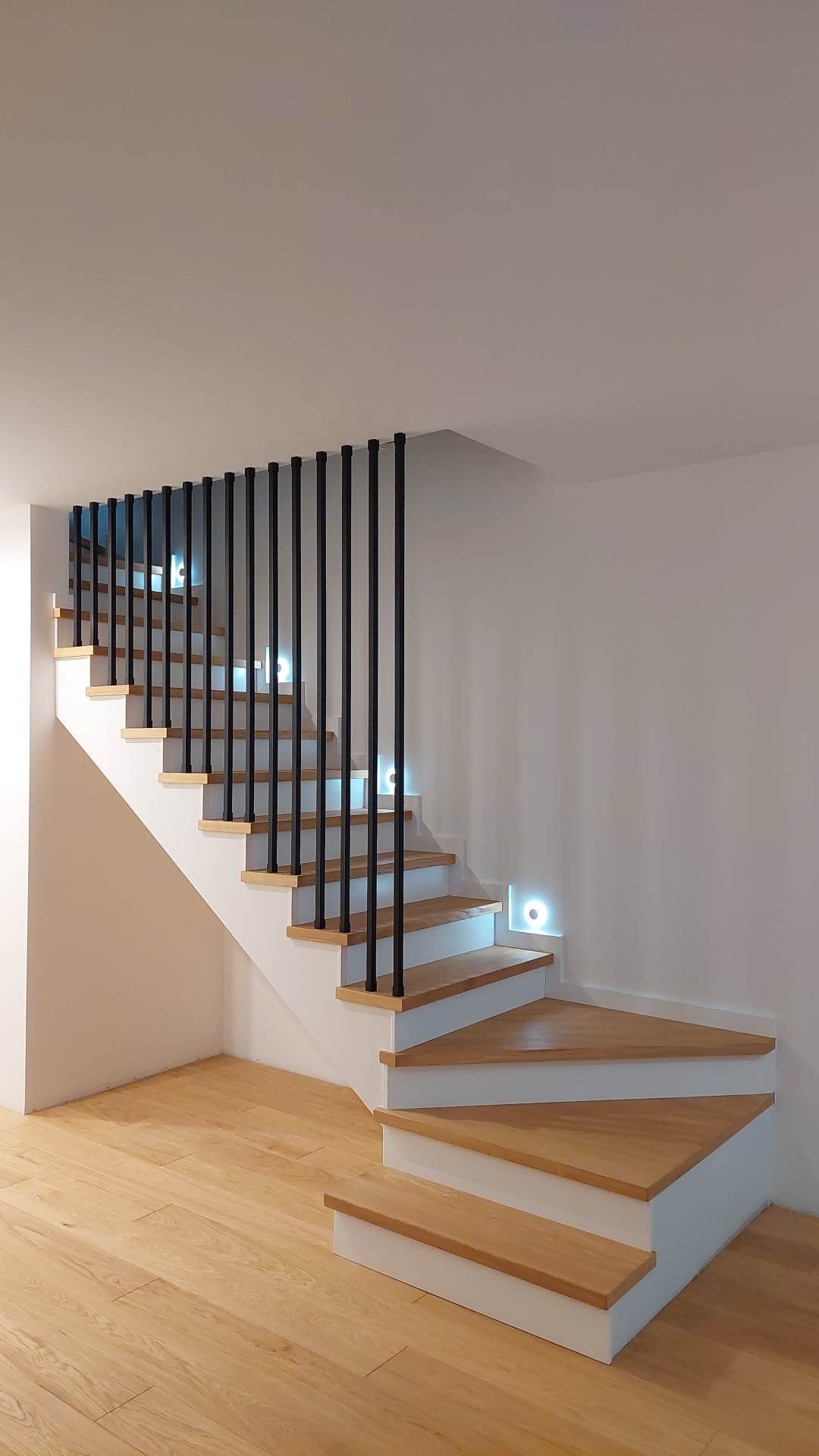 Nowoczesne schody drewniane z białymi stopnicami i czarną, minimalistyczną balustradą z okrągłych prętów, podświetlone punktowymi lampkami LED w ścianie.