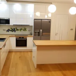 Nowoczesna, biała kuchnia z drewnianym blatem, wyspą i zabudową pod sufit, oświetlona kulistymi lampami wiszącymi.