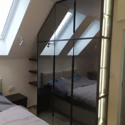 Szafa wnękowa z lustrzanymi drzwiami na czarnym stelażu, z oświetleniem LED, odbijająca wnętrze sypialni na poddaszu z oknami dachowymi.