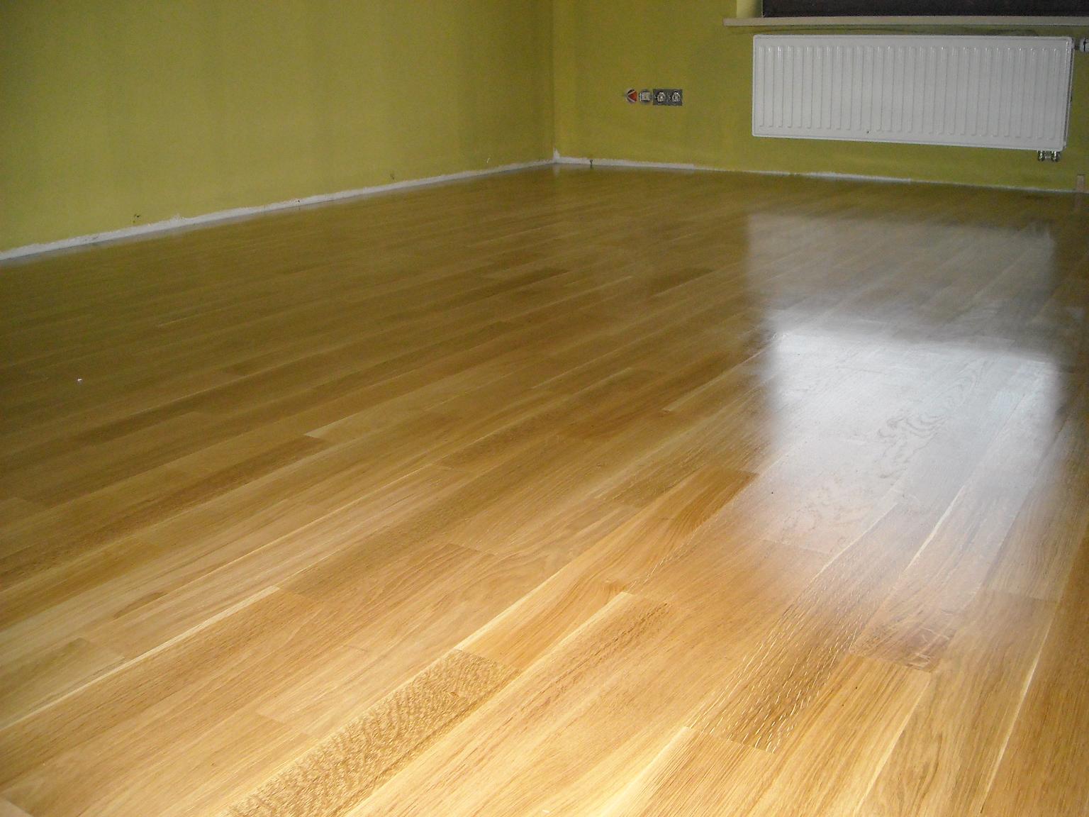 Wycyklinowana, lakierowana podłoga parkietowa z widocznym odbiciem światła w pustym pokoju z żółtymi ścianami i białym grzejnikiem.