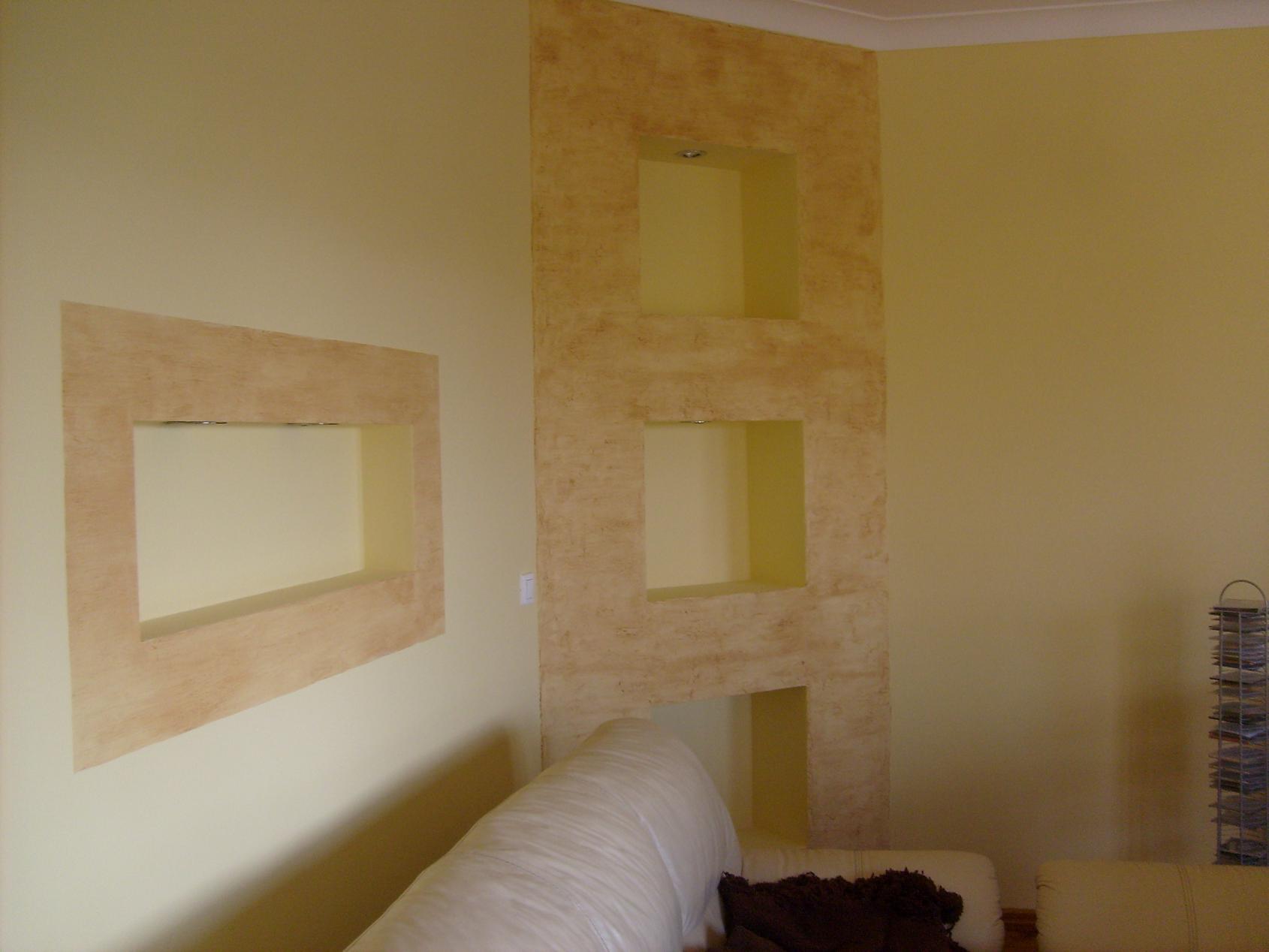 Wykończona ściana z wnękami, pokryta dekoracyjną strukturą w odcieniach beżu i brązu, widoczna z perspektywy narożnika pokoju, z fragmentem sofy w dolnej części kadru.