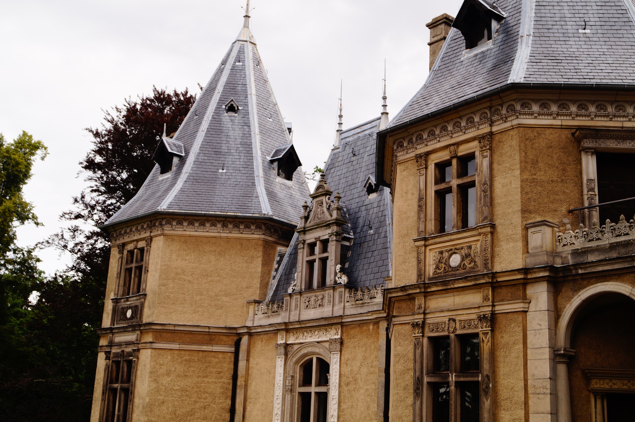 Zewnętrzny widok na fragment zabytkowego pałacu z wieżyczkami, szarą dachówką i dekoracyjnymi oknami w stylu neogotyckim.