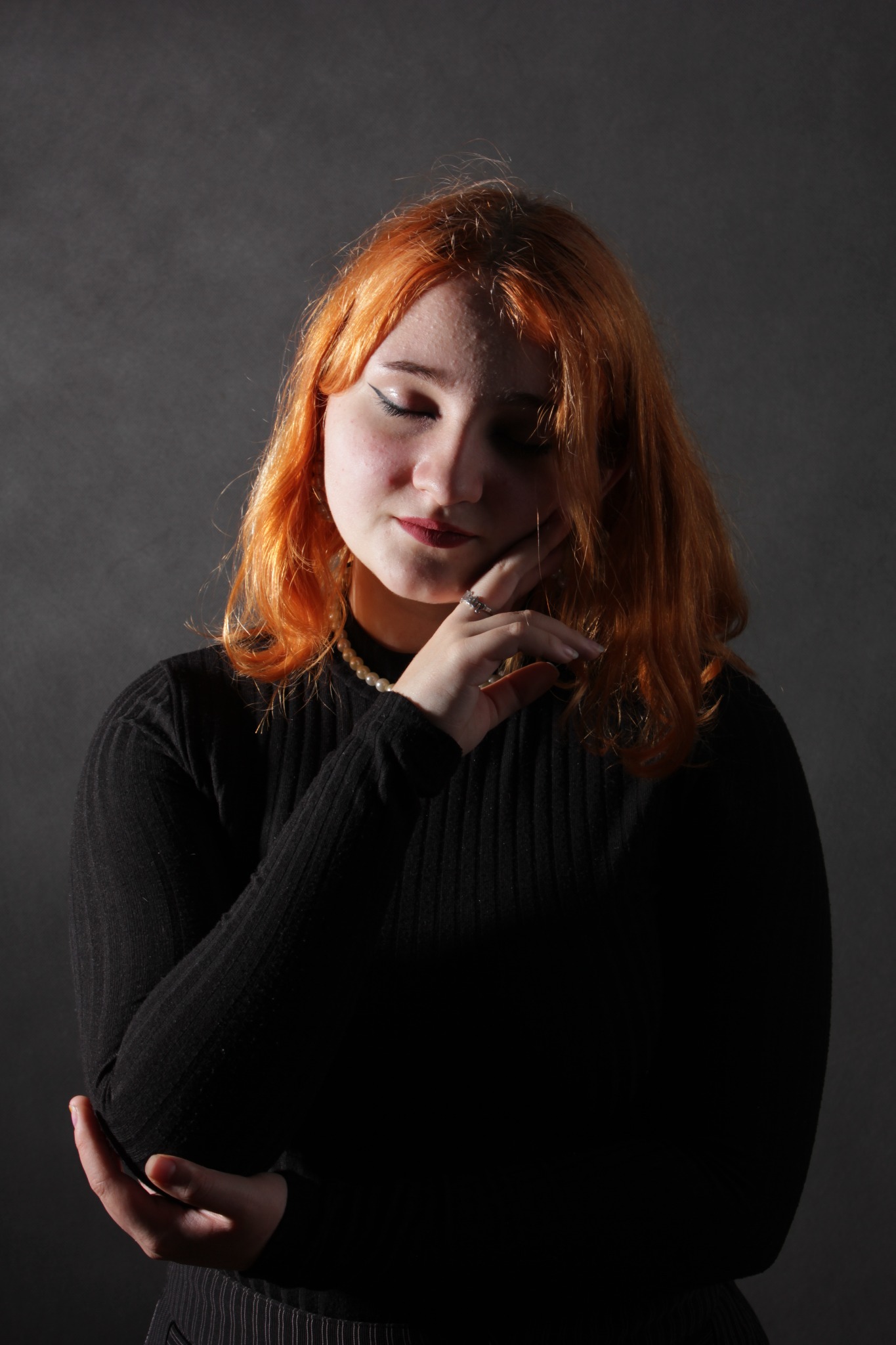 Portret kobiety z rudymi włosami w czarnym swetrze, z delikatnym makijażem i perłowym naszyjnikiem, pozującej w Konarzewie na ciemnoszarym tle.