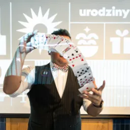 Mężczyzna w muszce prezentuje trik z wachlarzem kart na tle projekcji z napisem 'urodziny' i grafikami prezentów.