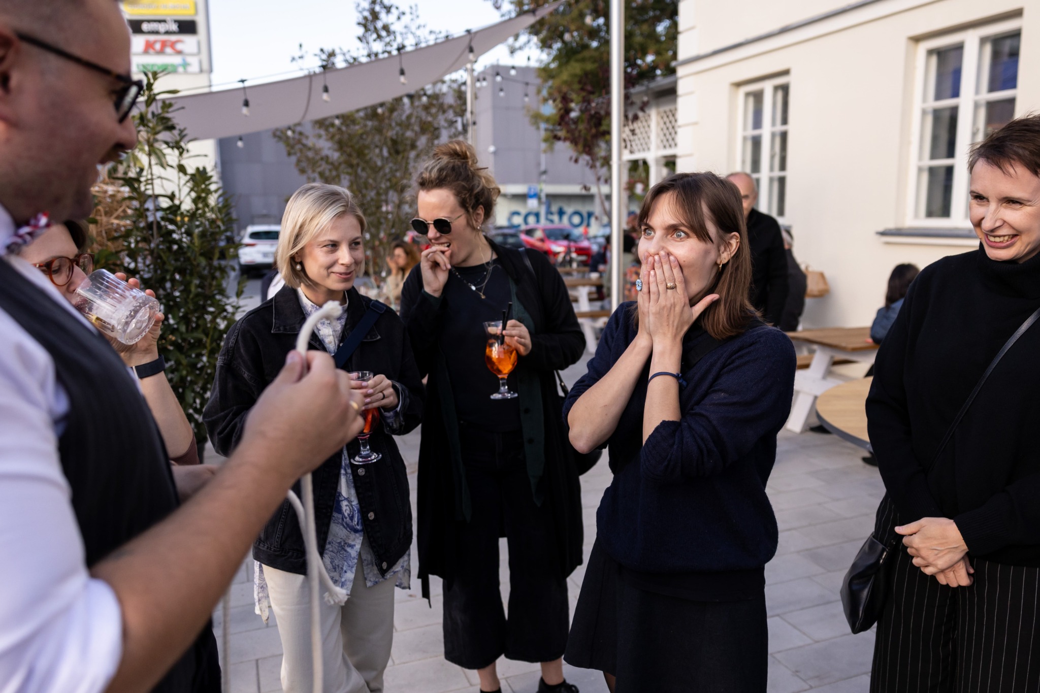 Magik prezentuje trik z liną przed grupą zaskoczonych kobiet na zewnątrz budynku z markizami i oświetleniem.