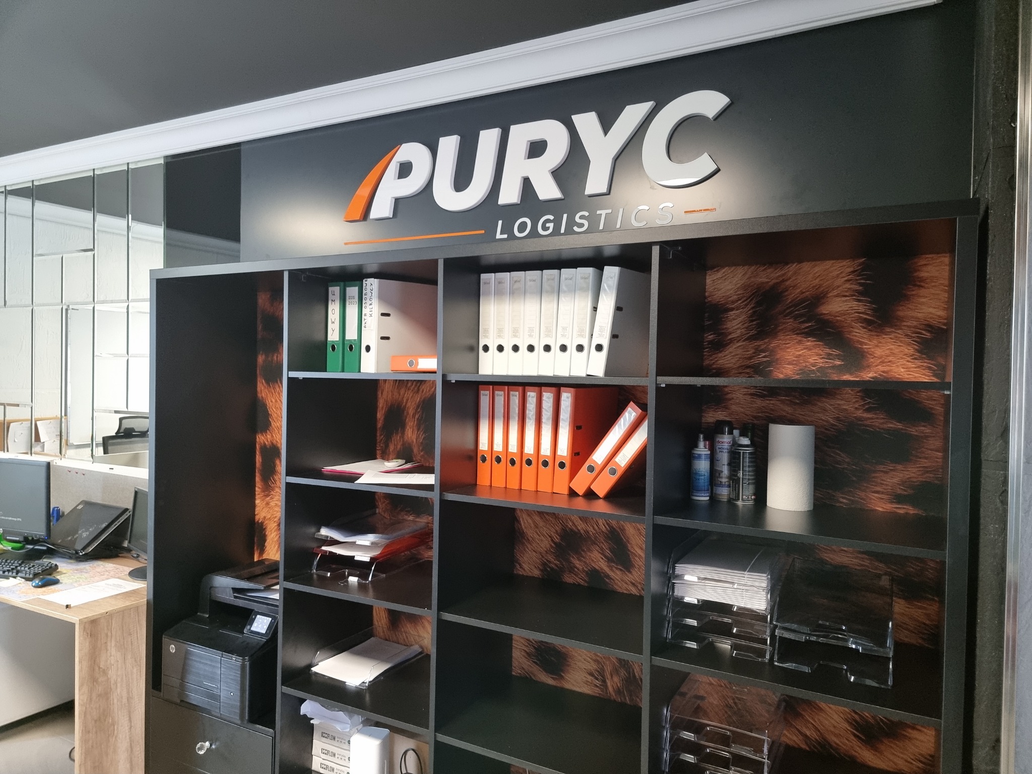 Biuro firmy logistycznej z regałem wypełnionym segregatorami i dokumentami, widoczne logo PURYC Logistics nad regałem.
