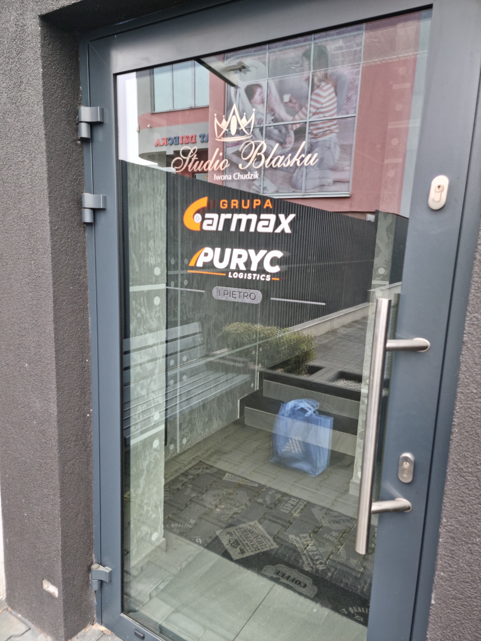 Szklane drzwi wejściowe do budynku z widocznymi logotypami firm Carmax i Puryc Logistics oraz Studio Blasku, na wycieraczce napis Espresso Coffee Quality