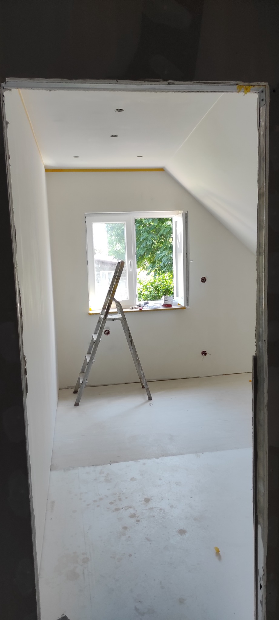 Pokój w trakcie remontu: białe ściany po gładzi, zabezpieczone taśmą malarską przy suficie, drabina aluminiowa przy oknie, podłoga z widocznymi zabrudzeniami po remoncie, widok przez ościeżnicę.