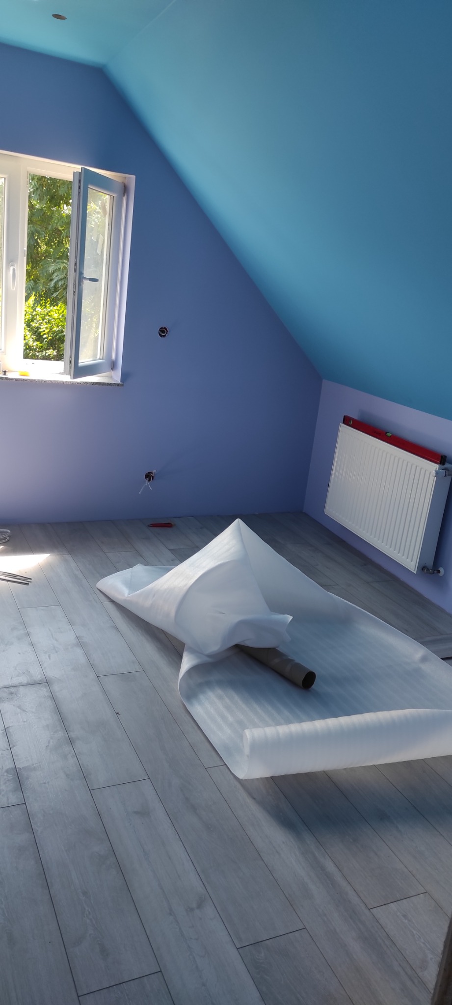 Pokój na poddaszu z niebieskimi ścianami i panelami podłogowymi w trakcie prac wykończeniowych, zabezpieczone panele folią ochronną, widoczny grzejnik i okno.