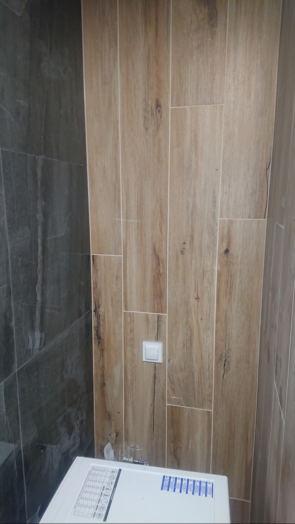 Fragment łazienki z częściowo ułożonymi płytkami imitującymi drewno na ścianie, widoczny biały przycisk spłuczki i fragment pralki.